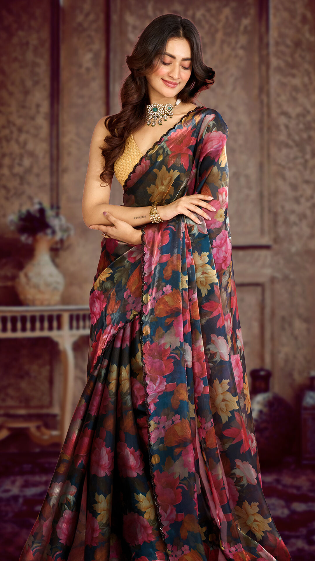 Elegant woman in a black floral printed saree with pink and orange blossoms, paired with a golden blouse and statement choker necklace, posing gracefully in a vintage-inspired indoor setting.